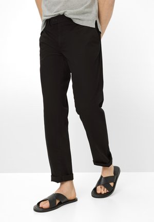 Man met zwarte opgerolde broek en zwarte sandalen met kruisbanden, staand met handen achter de rug tegen een witte achtergrond.