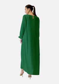 Długa zielona sukienka z długimi rękawami, z otworem w kształcie klucza z tyłu, wykonana z gładkiego materiału, z ozdobnymi marszczonymi mankietami.