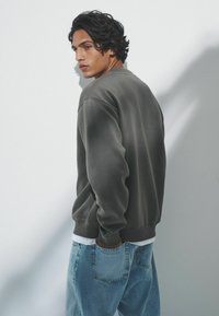 Sivá mikina vyrobená z mäkkej tkaniny, s kruhovým výstrihom a rebrovanými manžetami, skombinovaná s svetlomodrými džínami a bielou vrstvenou košeľou pod ňou.