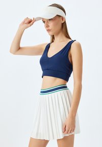 Navy blue urheilullinen crop-top, jossa on leveät hihnat, yhdistettynä pleatattuun valkoiseen hameeseen, jossa on monivärinen vyötärönauha. Valkoinen visori-asu.