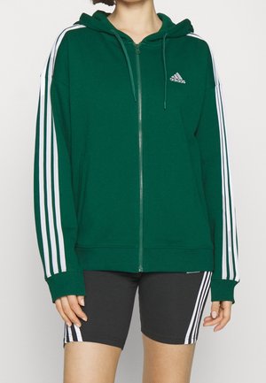 Persoon draagt een groen Adidas zip-up hoodie met witte strepen op de mouwen en zwarte Adidas shorts met witte strepen.