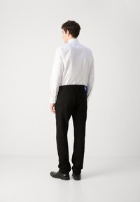 Jean en denim noir avec une coupe droite, arborant une étiquette bleue au niveau de la taille, associé à une chemise blanche à manches longues et des chaussures noires.