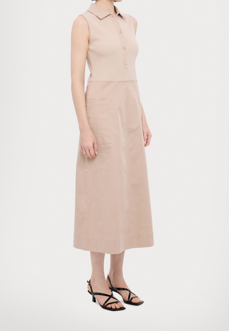 Robe midi beige sans manches avec col et patte de boutonnage, portée avec des sandales à talons noires à brides, sur une personne debout.