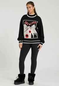 Zwarte gebreide trui met een "LES ALPES" graphic, witte accenten en een comfortabele textuur. Draagt met zwarte leggings en harige zwarte laarzen.