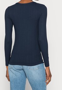 Person klädd i en åtsittande marinblå ribbad långärmad skjorta och ljusblå denimjeans, sedd bakifrån mot en enfärgad bakgrund.