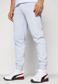 Light gray sweatpants made of soft fabric with a fitted design. Black logo detail on the thigh and ribbed cuffs. Worn with white sneakers.