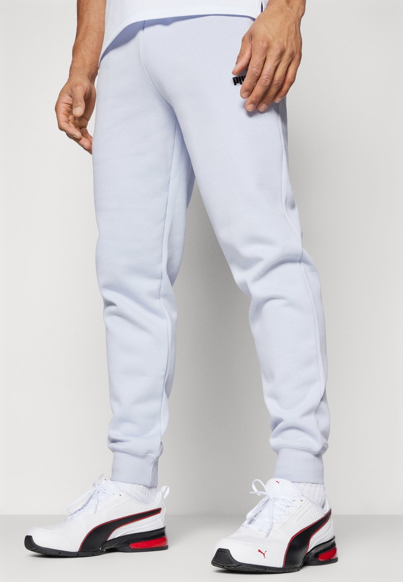 Pantalones de chándal gris claro, hechos de una tela suave y con un diseño ajustado. Detalle del logo negro en el muslo y puños acanalados. Llevados con zapatillas blancas.