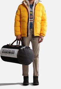 Yellow puffer jacket with a zip closure, beige pants, black boots, and a round black and white duffel bag with branding.