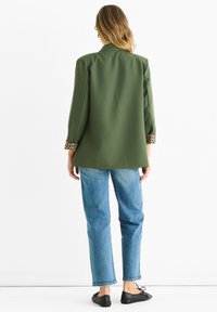 Blazer verde oliva con polsini in stampa leopardata, abbinato a jeans di denim azzurro chiaro. Texture liscia e design su misura, con uno spacco sul retro.