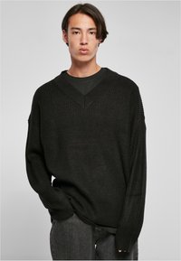 Suéter negro de punto extragrande con un escote en forma de V profundo, textura acanalada y hombros caídos, superpuesto a una camiseta negra.