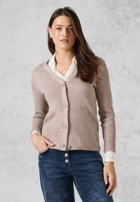 Hellbrauner, gerippter Cardigan mit V-Ausschnitt, Knopfverschluss, langen Ärmeln und über einem weißen Hemd mit Kragen getragen. Sichtbare Jeans aus Denim darunter.