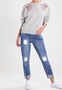 Felpa grigia con ricami floreali sulle spalle, abbinata a jeans blu strappati con orli arrotolati e sneakers bianche.