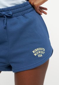 Μπλε σορτς με κορδόνι που φοριούνται από άτομο, με κίτρινο κείμενο "MINNESOTA MIDWEST USA" στο μπροστινό αριστερό μηρό.