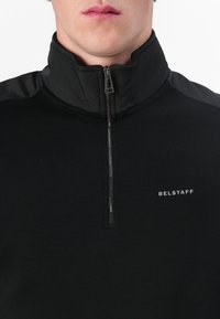 Čierny pulóver s vysokým golierom, polovičným zipsom a jemnou textúrou. Na hrudi má diskrétnu nášivku "Belstaff".