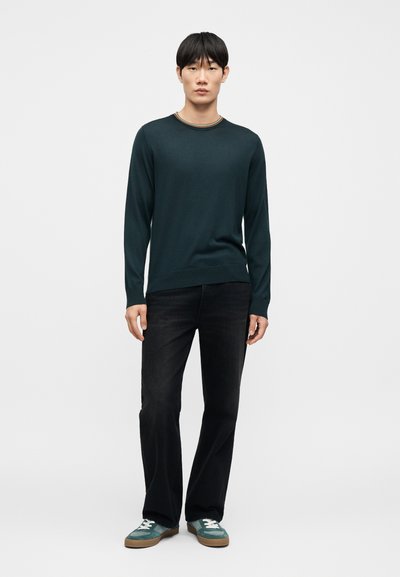 Pull vert foncé à manches longues avec un col côtelé orné d'une fine bande multicolore, assorti à un jean noir et des baskets bleu sarcelle.