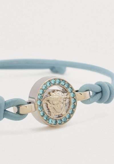 Bracelet en cuir bleu clair avec une fermeture ronde en ton argent. La fermeture présente une tête de lion en relief entourée de strass turquoise.