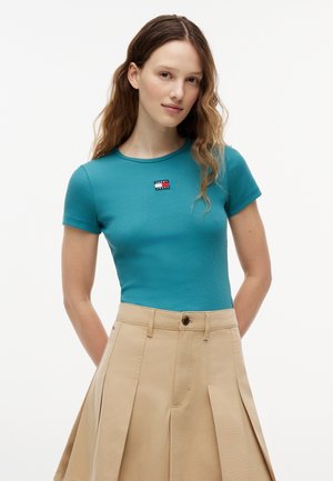 Jeune femme aux longs cheveux ondulés portant un haut ajusté à manches courtes bleu sarcelle et une jupe plissée taille haute beige, debout devant un fond blanc.