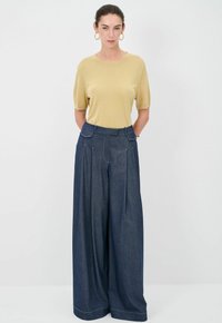 Top a maniche corte giallo chiaro abbinato a pantaloni larghi in denim scuro; i pantaloni presentano pince frontali e dettagli di cucitura a contrasto.