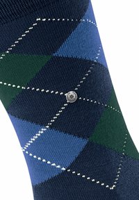 Acercamiento a una media argyle de color azul, verde y marino con un patrón de diamantes bordado en blanco y un pequeño botón de metal redondo con logotipo.