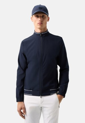 Mand iført marineblå lynlåsjakke med hvid kant, matchende marineblå kasket og hvide bukser, stående med den ene hånd i lommen.