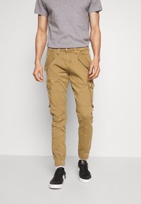 Pantalones cargo caqui con múltiples bolsillos, detalles de cremallera y corte ajustado. Combinados con zapatillas negras. Diseño sencillo y utilitario.