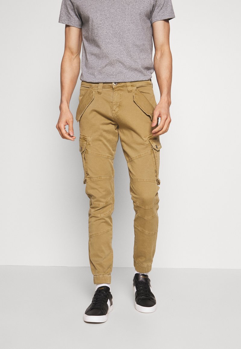 Pantalones cargo caqui con múltiples bolsillos, detalles de cremallera y corte ajustado. Combinados con zapatillas negras. Diseño sencillo y utilitario.