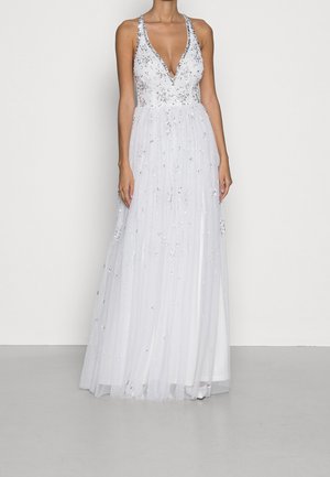 Mouwloze witte jurk met diepe V-hals, zilveren bloemrijke versieringen en een vloeiende tule rok, gedragen door een slanke staande vrouw.