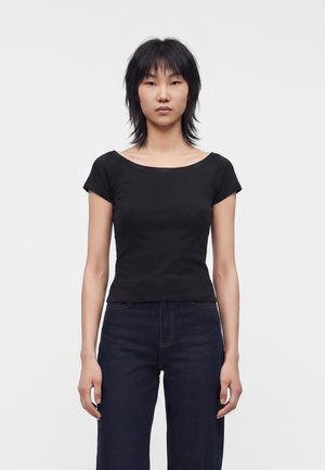 Jeune personne aux cheveux noirs courts portant un haut noir ajusté à manches courtes et un jean foncé taille haute, debout devant un fond uni.