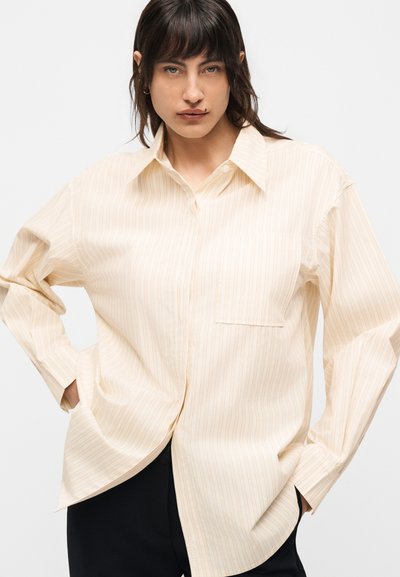 Άτομο που φοράει ένα φαρδύ, ανοιχτό μπεζ ριγέ πουκάμισο με γιακά και μακριά μανίκια, τα χέρια στις τσέπες, κοιτάει μπροστά σε απλό φόντο.