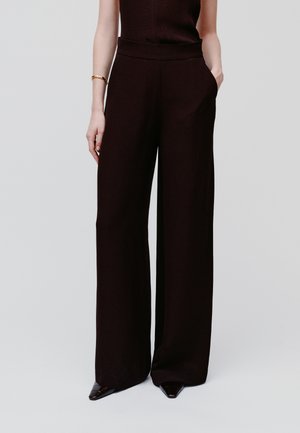 Femme portant un pantalon noir taille haute à jambes larges, avec une main dans la poche, associé à des chaussures noires à talons pointus et un bracelet en or au poignet.