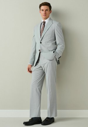 Young man standing against wall wearing light gray suit, patterned tie, white shirt, and black loafers with hand in pocket.