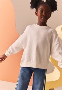 Felpa oversize color avorio con polsini e orlo a coste, abbinata a jeans blu con strisce laterali verdi. Tessuto liscio, design casual.