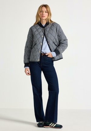 Kvinde med blondt hår iført en sort og hvid ternet quiltet jakke, lyseblå sweater, mørkeblå jeans og sorte sneakers med hvide striber.