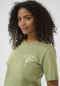 T-shirt en coton vert olive avec un texte graphique blanc et une illustration de cocktail sur le côté gauche de la poitrine, à manches courtes et à coupe décontractée.