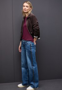 Brązowa skórzana kurtka bomberka z białymi i czarnymi paskami na mankietach, fioletowa koszula, szerokie niebieskie dżinsy z bocznymi paskami oraz białe sneakersy.