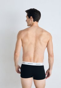 Boxers noirs en tissu avec une ceinture blanche portant le logo Lacoste. Modèle vu de dos avec des cheveux bruns courts et sans chemise.
