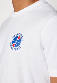 T-shirt en coton blanc avec un imprimé circulaire bleu, rouge et blanc portant les mots "BASKETBALL" et "Champion". Design classique à col rond.