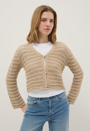 Mujer joven con cabello rubio hasta los hombros, vistiendo un cárdigan beige de crochet sobre una camisa blanca y jeans azules, de pie con las manos en las caderas.