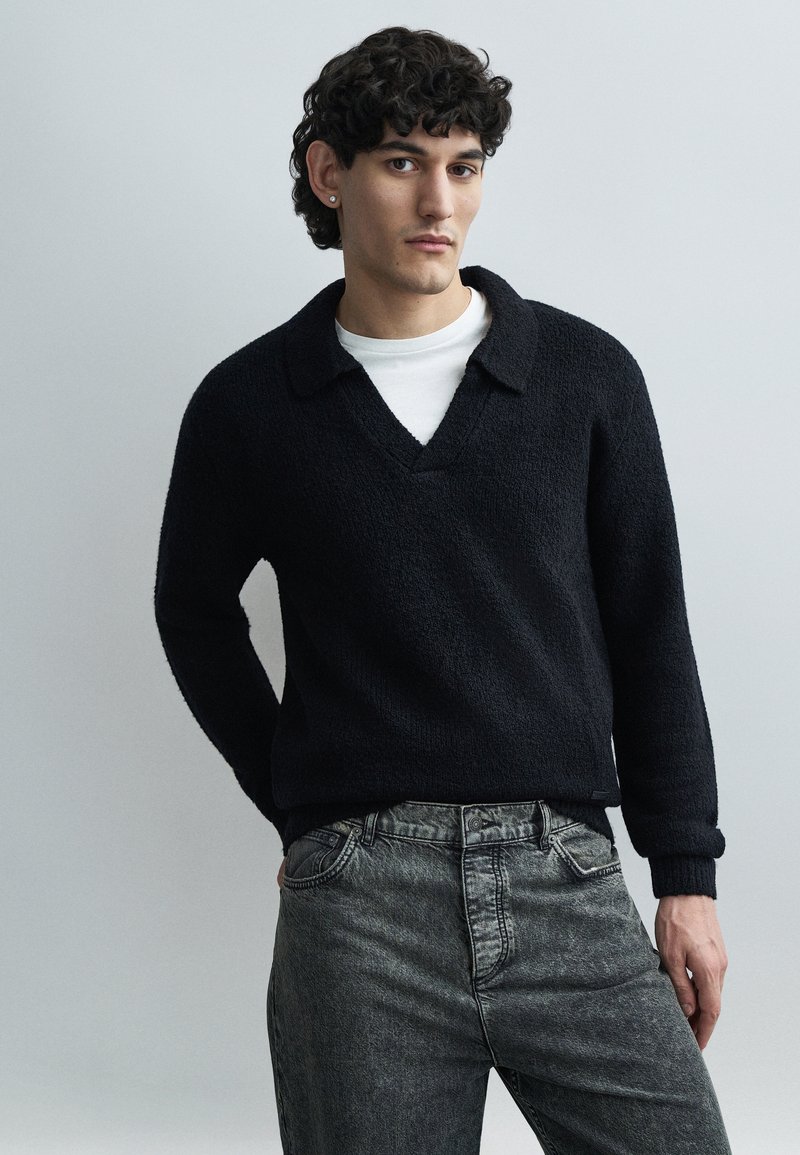 Tânăr cu păr creț și întunecat, purtând un pulover negru texturat peste o cămașă albă și blugi negri decolorați, stând în fața unui fundal simplu și deschis la culoare.