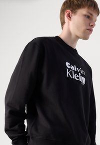 Black sweatshirt made of cotton with a ribbed collar. Features white "Calvin Klein" logo printed on the front. Soft texture and relaxed fit.
