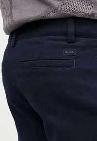 Gros plan sur un pantalon bleu marine avec une poche passepoilée à l’arrière et une petite étiquette de marque BOSS au-dessus de la poche, porté avec un pull beige texturé.