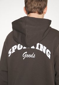 Personne portant un sweat à capuche foncé avec les mots "SPORTING Goods" imprimés en blanc dans le dos, vue de dos.