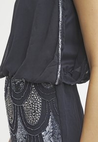 Haut en tissu marine avec des manches transparentes et un travail de perles complexe en motif ondulé sur la moitié inférieure, créant une apparence texturée.