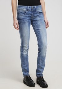 Jeans de mezclilla azul con corte slim, que presentan una cintura de altura media, cinco bolsillos, un ligero desgaste y detalles de costura. Combinados con calzado negro.