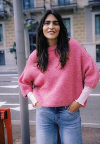 Pull rose surdimensionné en tissu duveteux avec poignets côtelés, superposé sur un haut à manches longues blanc ; associé à un jean bleu clair. Environnement urbain en extérieur.