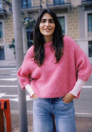 Lielizmēra rozā pūkains džemperis ar ribotām aprocēm, kārtots pār baltu garām piedurknēm topu; kombinēts ar gaiši zilām džinsu biksēm. Pilsētas āra vide.