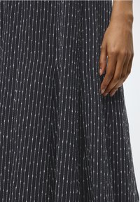 Το δεξί χέρι ακουμπά δίπλα σε μια μαύρη φούστα με λευκές κάθετες διακεκομμένες ρίγες σε μια επίπεδη, φωτεινή επιφάνεια.