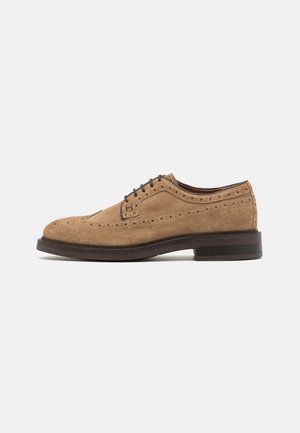 Chaussure habillée brogue en daim beige avec détails perforés, lacets noirs et semelle marron foncé, vue de côté extérieur sur fond blanc.