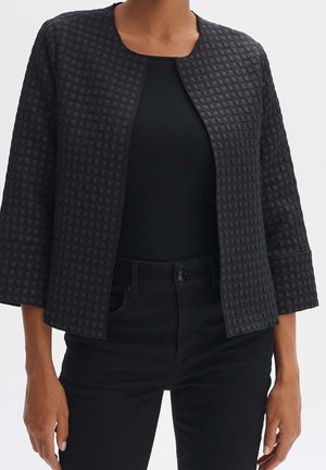 Chaqueta negra acolchada con patrón de cuadros, abierta por delante, con mangas tres cuartos, llevada sobre una camiseta negra y pantalones negros.