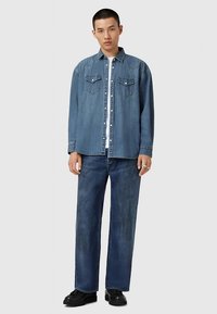 Camicia di denim e pantaloni a gamba larga in blu medio e scuro. La camicia presenta due tasche sul petto e chiusura frontale con bottoni. Le scarpe sono nere con una suola spessa.